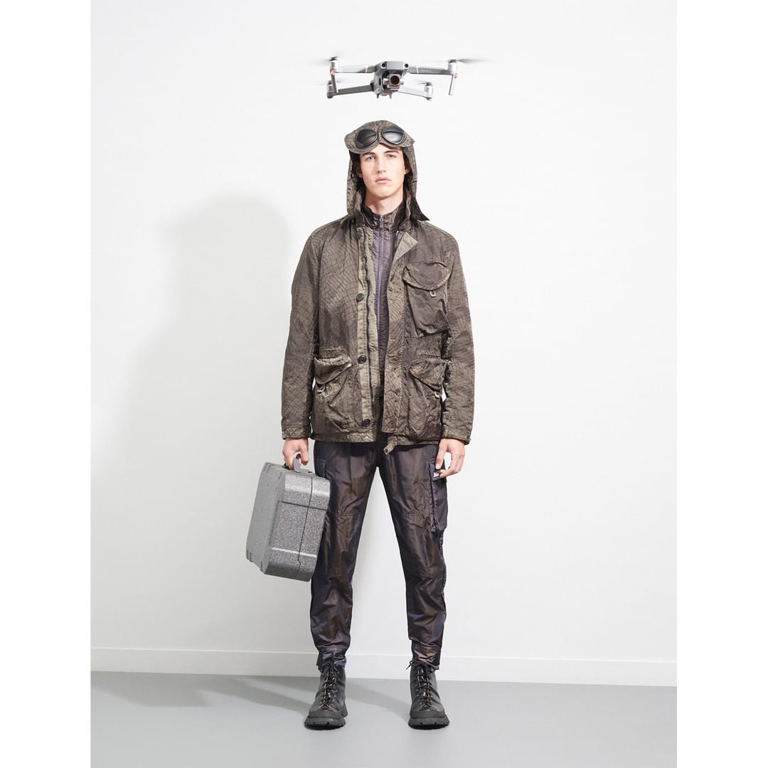 Adult male in aviator cap and olive jacket stands indoors holding gray toolbox, drone flies overhead, against white wall. Fashion shot.