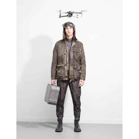 Adult male in aviator cap and olive jacket stands indoors holding gray toolbox, drone flies overhead, against white wall. Fashion shot.