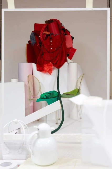 Artistic display featuring a large flower sculpture made of red fabric and clothing, set against a white backdrop with transparent chairs.