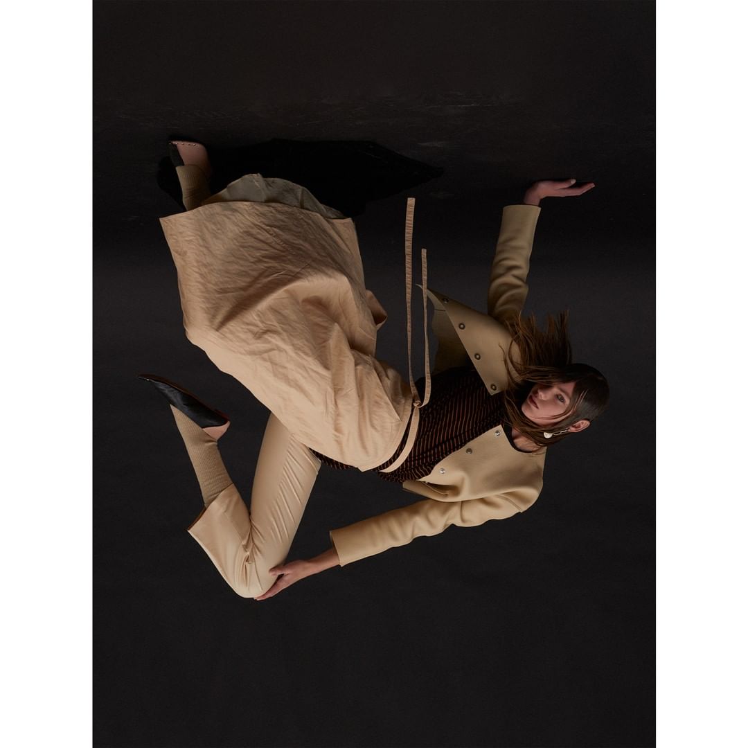A woman with long hair poses in a beige jacket, trousers, and skirt against a dark background, showcasing a fashion-forward look.