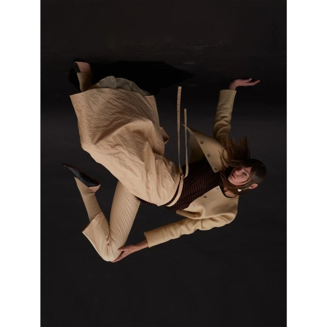 A woman with long hair poses in a beige jacket, trousers, and skirt against a dark background, showcasing a fashion-forward look.