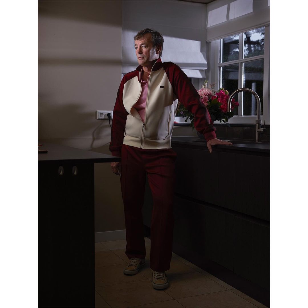 Mature adult male in a burgundy tracksuit stands in a modern kitchen with dark countertops and white cabinets, looking toward the window.