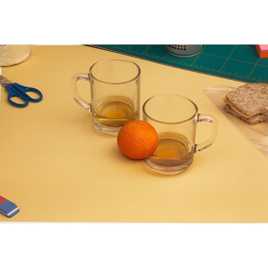 Still life of two clear glass mugs filled with yellow liquid, an orange, sandwich, scissors and other items on a yellow surface.