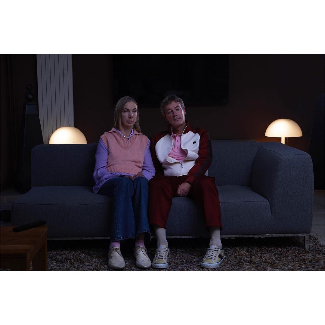 Indoor shot of a mature couple sitting on a blue sofa in front of a dark wall, lit by table lamps on either side, looking forward.