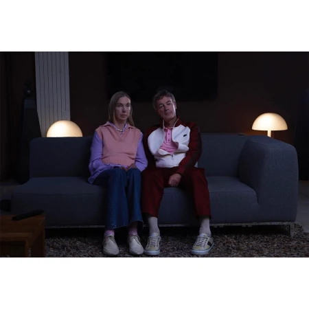 Indoor shot of a mature couple sitting on a blue sofa in front of a dark wall, lit by table lamps on either side, looking forward.