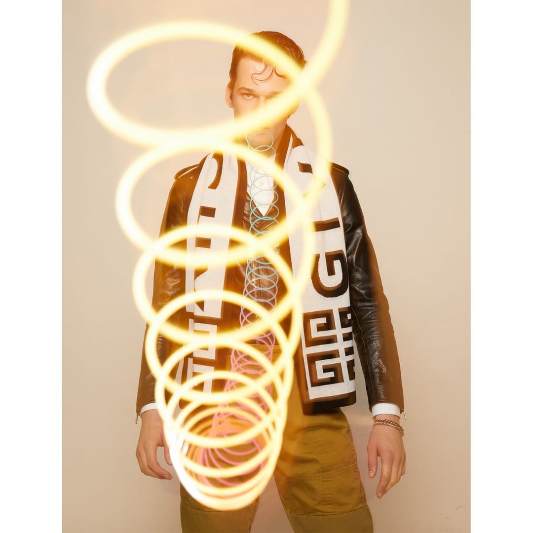 A fair-skinned male model in a black jacket, olive pants, and logo scarf poses against a light background with a glowing light effect.