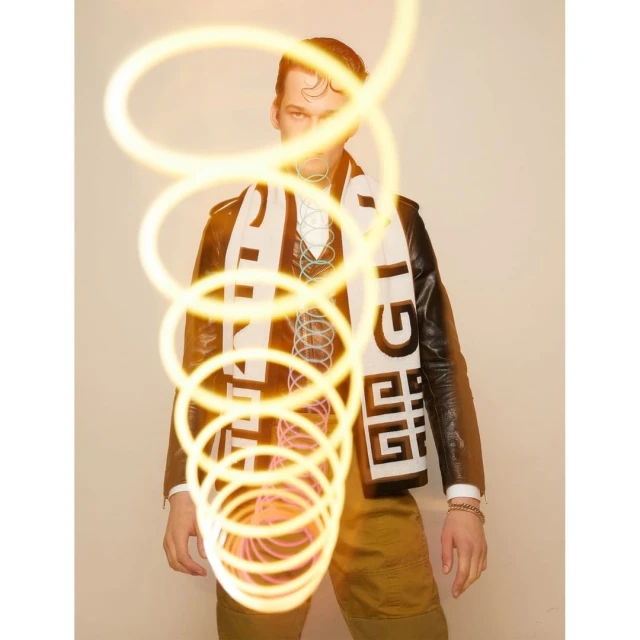 A fair-skinned male model in a black jacket, olive pants, and logo scarf poses against a light background with a glowing light effect.