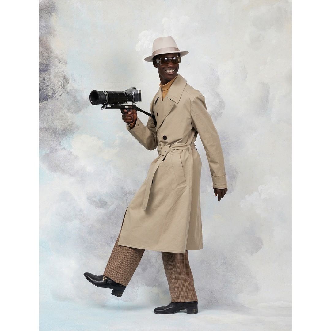 An adult male in a beige trench coat poses with a vintage camera in a studio with a cloudy backdrop, creating a film noir style image.