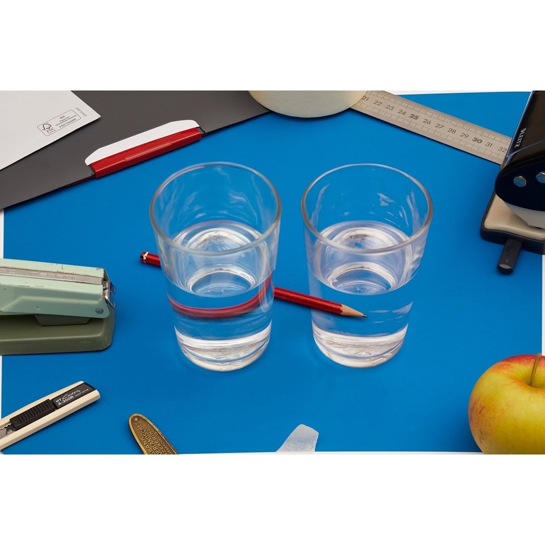 Two glasses of water with pencils on a blue surface among office supplies, including a stapler, ruler, apple and letter opener.