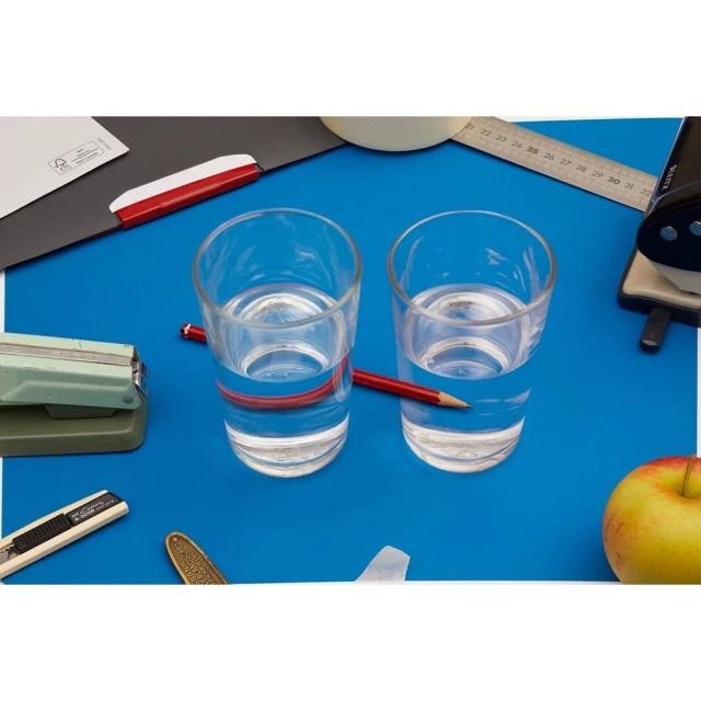 Two glasses of water with pencils on a blue surface among office supplies, including a stapler, ruler, apple and letter opener.