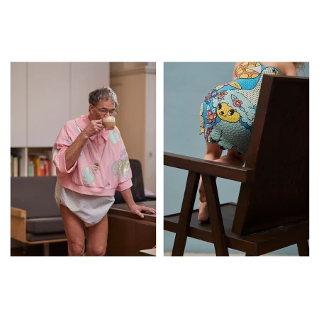 Older adult in pink top drinking coffee near sofa and bookshelves alongside a child with cartoon-printed bottoms near chair.