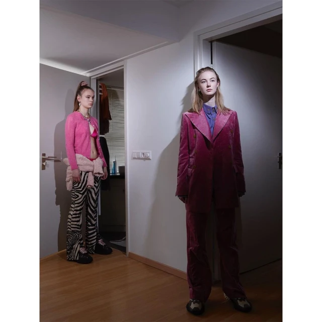 Two young women in stylish outfits pose indoors. One wears a pink top and zebra-print pants; the other, a pink velvet suit.