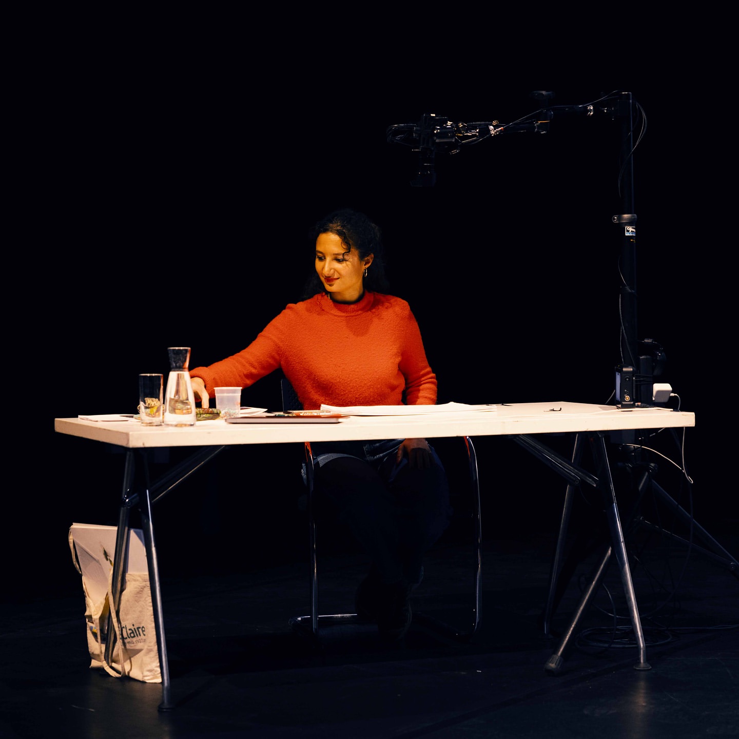 Woman in orange sweater sits at white table on stage with water jug and camera equipment, dark background. Stage setup, medium shot.