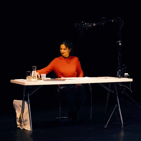 Woman in orange sweater sits at white table on stage with water jug and camera equipment, dark background. Stage setup, medium shot.