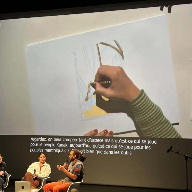 Close-up of a drawing on a screen with a hand using a crayon, with a panel discussion in the foreground.
