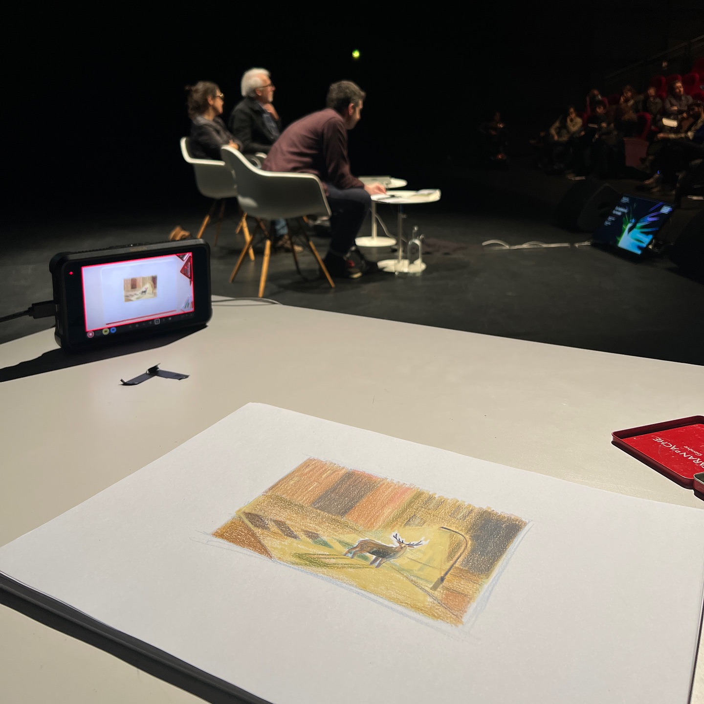 Illustration of a deer on a table in a dark room, likely part of a presentation with people in the background. Art, design, media.