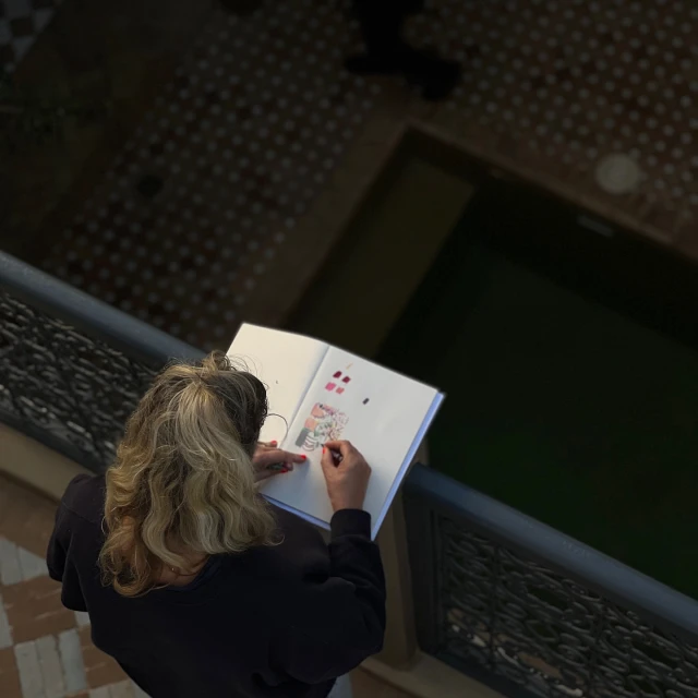 Woman sketches in an open notebook, viewed from above, with a patterned tiled floor and balcony visible in the background of the shot.