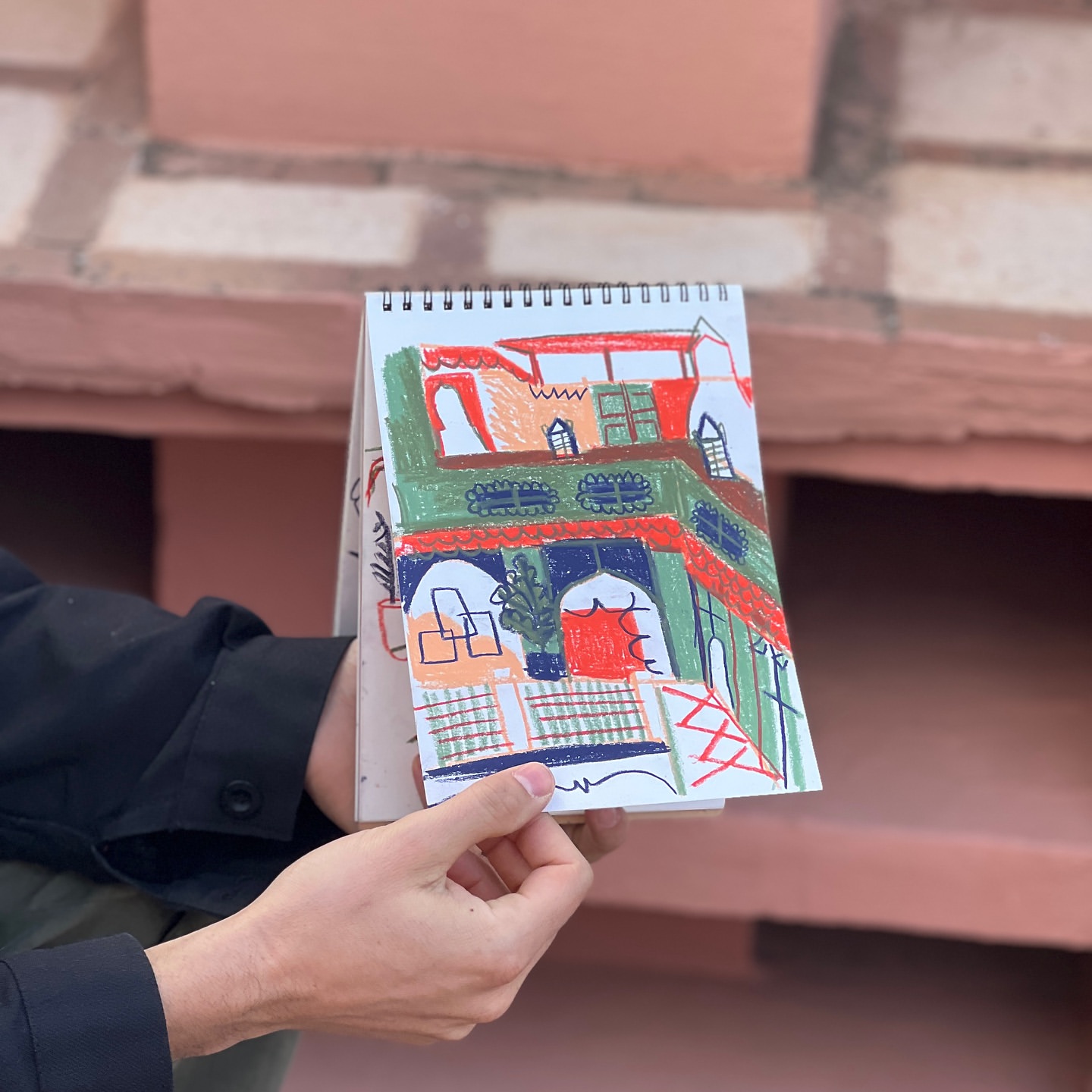 A hand holds open a sketchbook displaying a colorful drawing of a building with arches and windows, set against a brick wall backdrop.