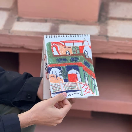 A hand holds open a sketchbook displaying a colorful drawing of a building with arches and windows, set against a brick wall backdrop.