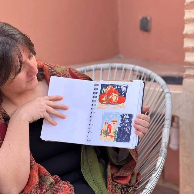 An adult woman sits in a white chair, holding open a sketchbook displaying vibrant tomato and building drawings, creating a cozy, artistic scene.