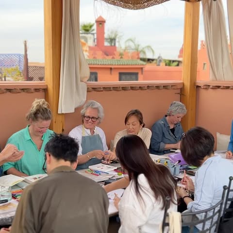 A group of people are gathered around tables on a rooftop, drawing or painting, under a partially covered area with red buildings behind.