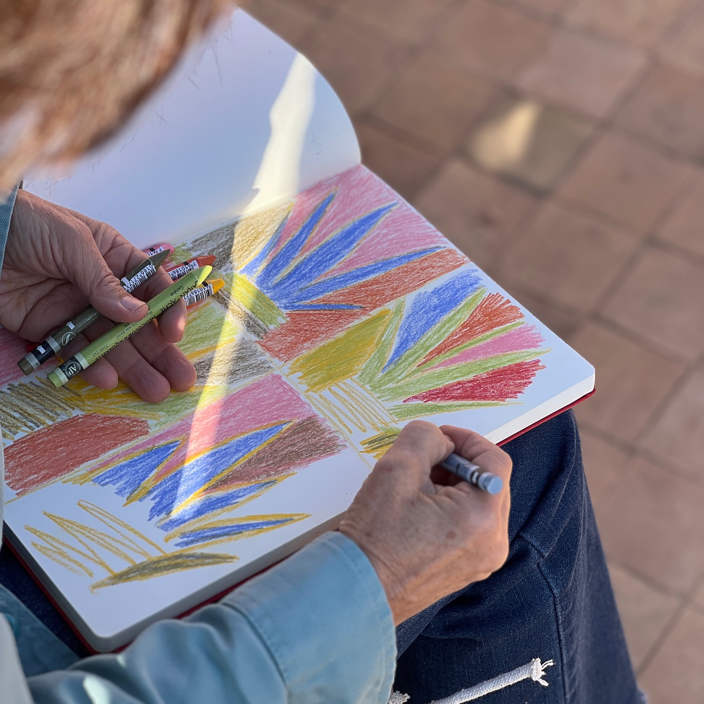 Woman artist creating colorful drawing in sketchbook with crayons, outdoor setting, close-up perspective, sunny daylight, casual wear.