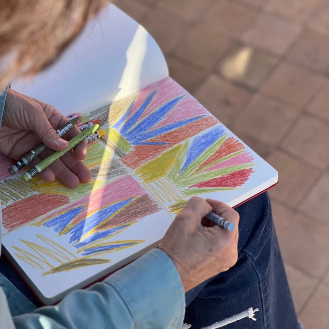 Woman artist creating colorful drawing in sketchbook with crayons, outdoor setting, close-up perspective, sunny daylight, casual wear.