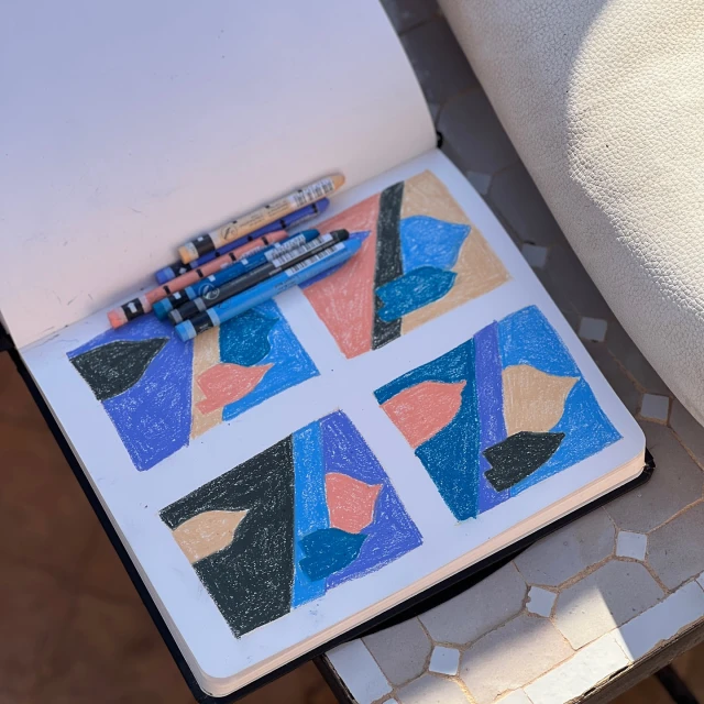 Open sketchbook featuring abstract crayon art on a mosaic table next to a neutral chair, capturing a creative and colorful tabletop scene.