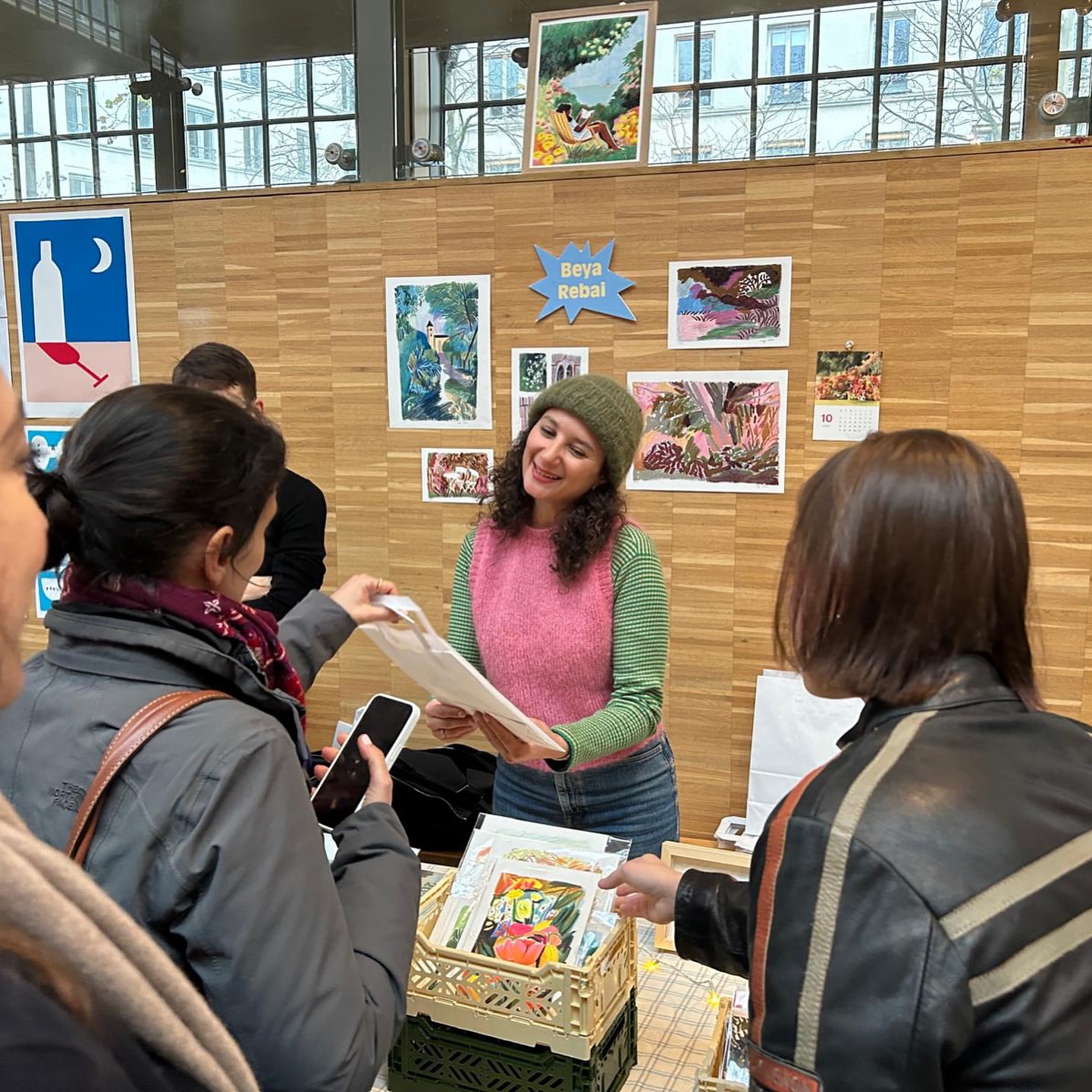 A woman with curly hair stands at an indoor market stall, selling art prints to customers, with natural light and a friendly atmosphere.