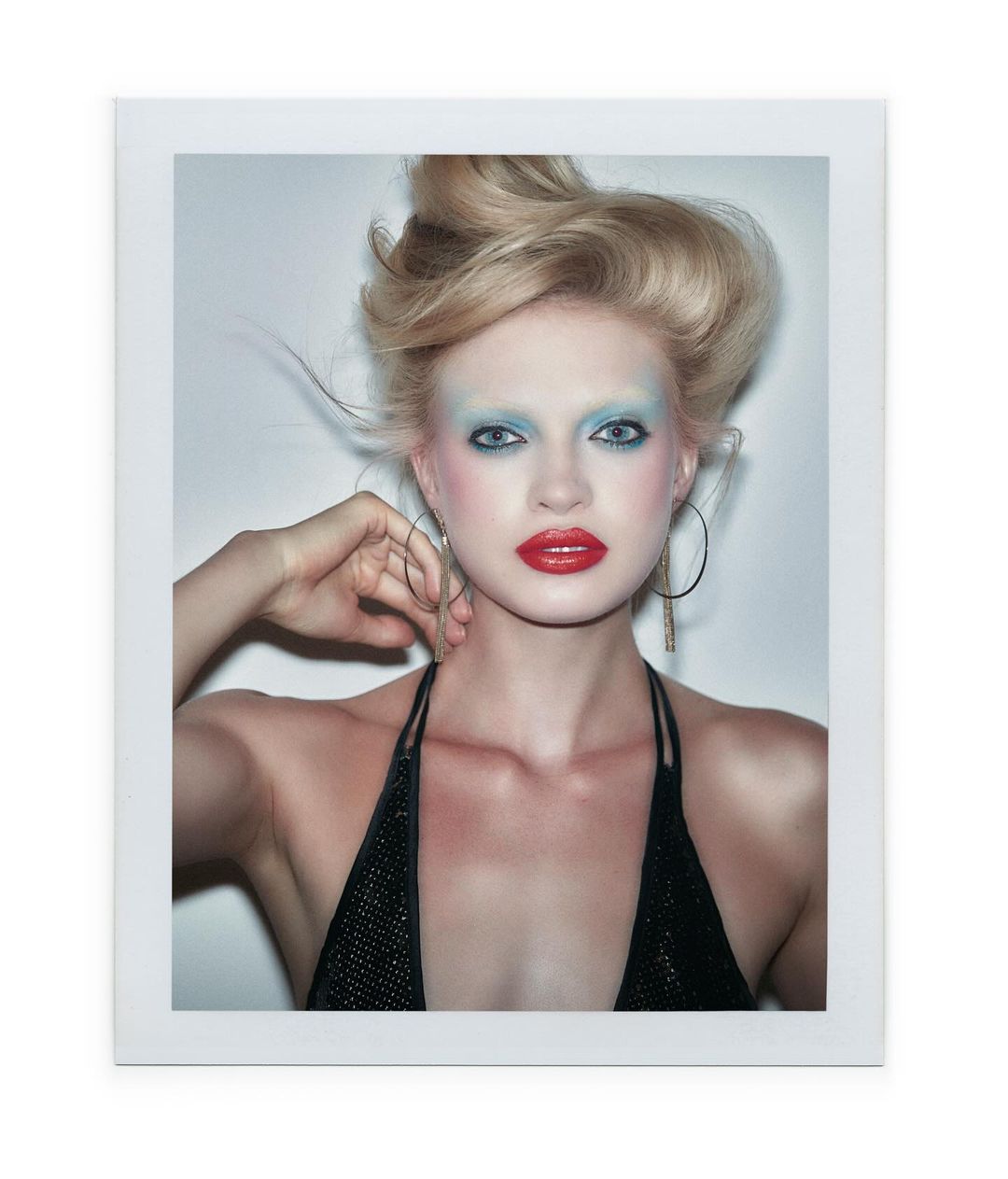 Close-up portrait of a blonde woman with blue eyeshadow, red lipstick, and large hoop earrings wearing a black top.