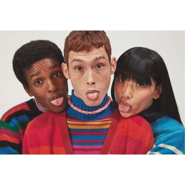 Three young adults playfully stick out their tongues in a light studio setting, wearing colorful striped sweaters for a fashion editorial.