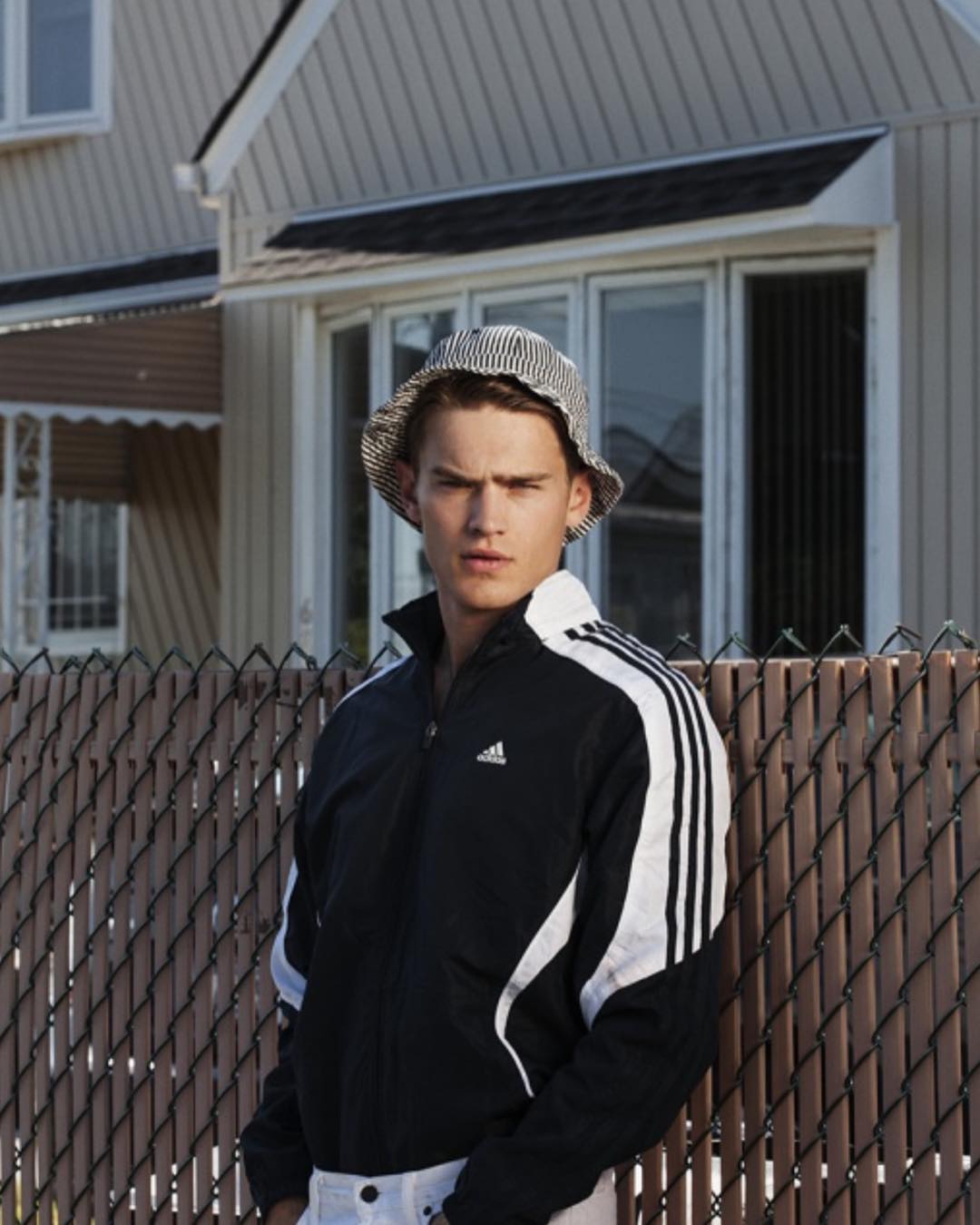 Adult male poses outdoors near fence wearing an Adidas track jacket and hat, medium shot. House is visible in the background.