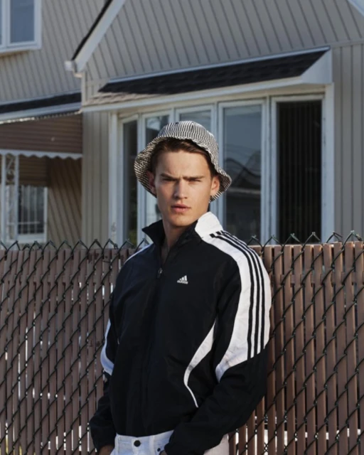 Adult male poses outdoors near fence wearing an Adidas track jacket and hat, medium shot. House is visible in the background.
