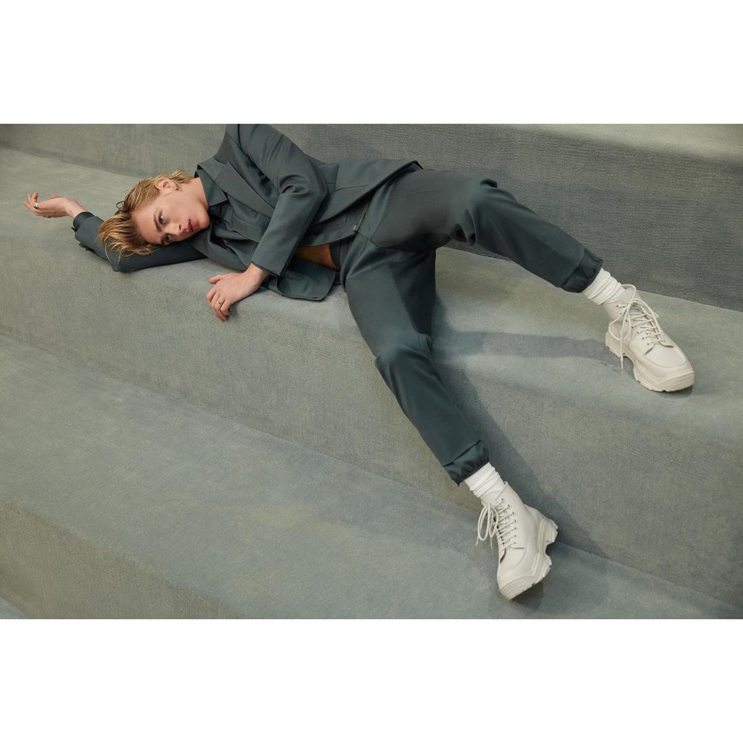 Woman in a teal suit and white platform sneakers lies on gray stairs in an indoor setting, showcasing a blend of fashion and casual style.