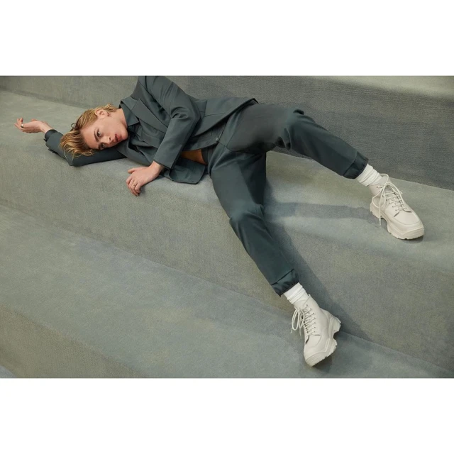 Woman in a teal suit and white platform sneakers lies on gray stairs in an indoor setting, showcasing a blend of fashion and casual style.