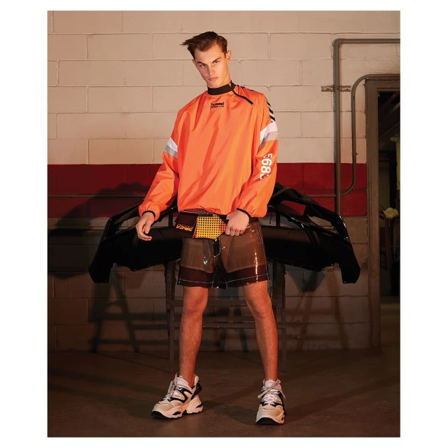Male fashion model in orange sweatshirt and transparent shorts poses in a garage with a car bumper in the background.