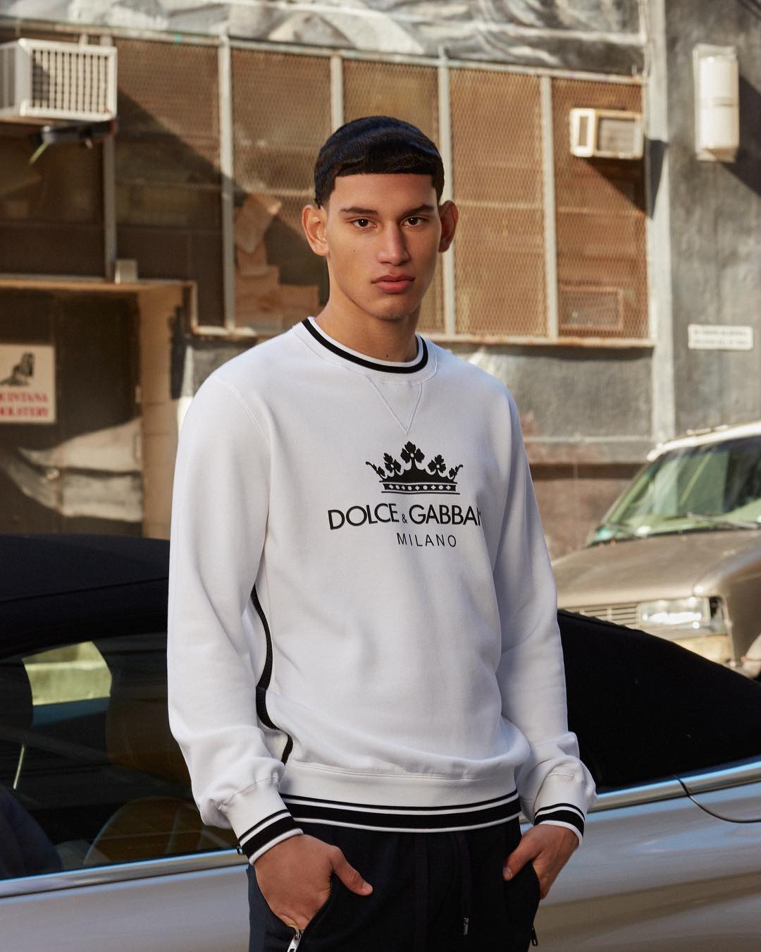 A stylish young man wearing Dolce & Gabbana sweatshirt and pants in an urban setting, leaning against a car, captured in natural light.