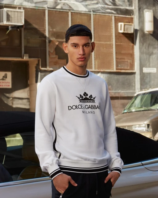 A stylish young man wearing Dolce & Gabbana sweatshirt and pants in an urban setting, leaning against a car, captured in natural light.