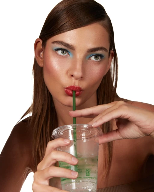 Attractive woman sips beverage through a green straw with vibrant blue eyeshadow and glossy red lips in a beauty-focused close-up studio shot.