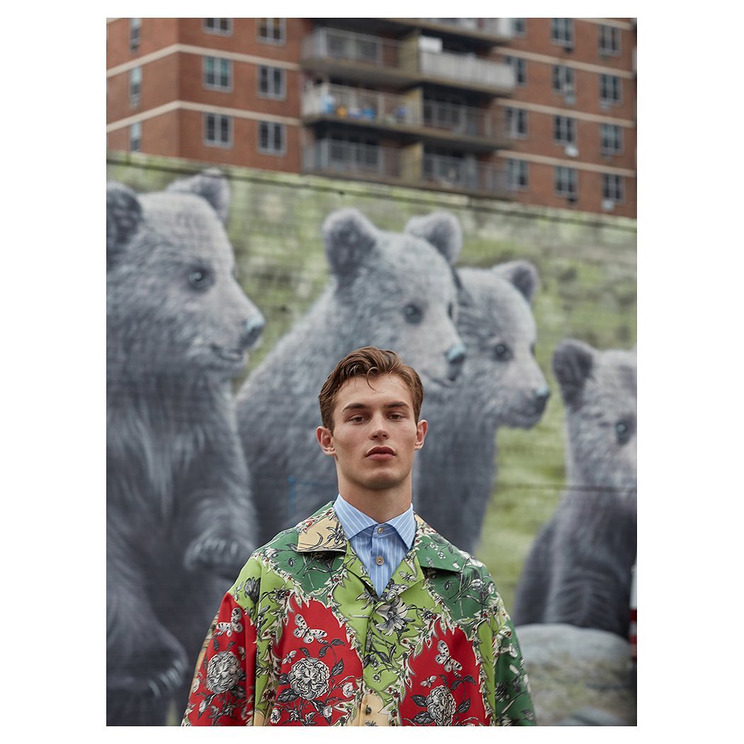 Male fashion model in patterned shirt poses in front of a mural of bear cubs in an urban setting with brick building in background.