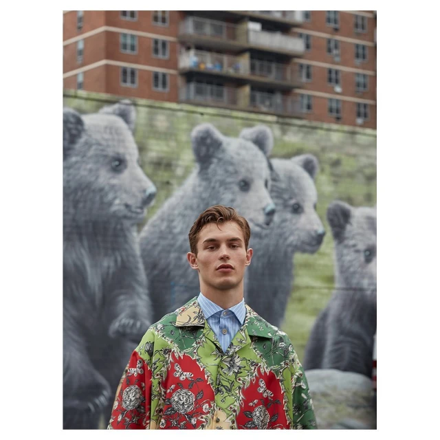 Male fashion model in patterned shirt poses in front of a mural of bear cubs in an urban setting with brick building in background.