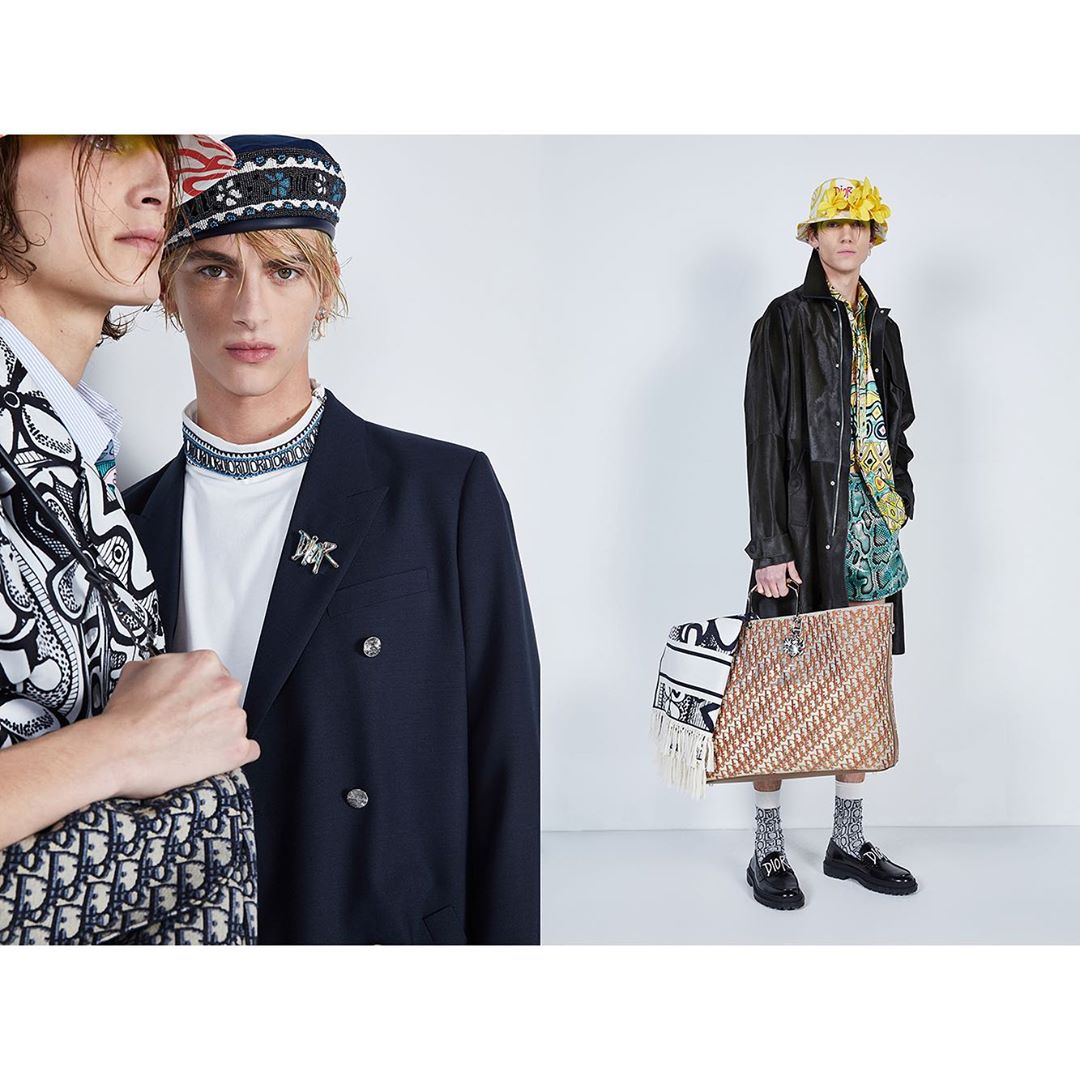 Two young adult fashion models pose in a photo studio wearing designer blazers, bucket hats, and carrying logo printed bags and accessories.