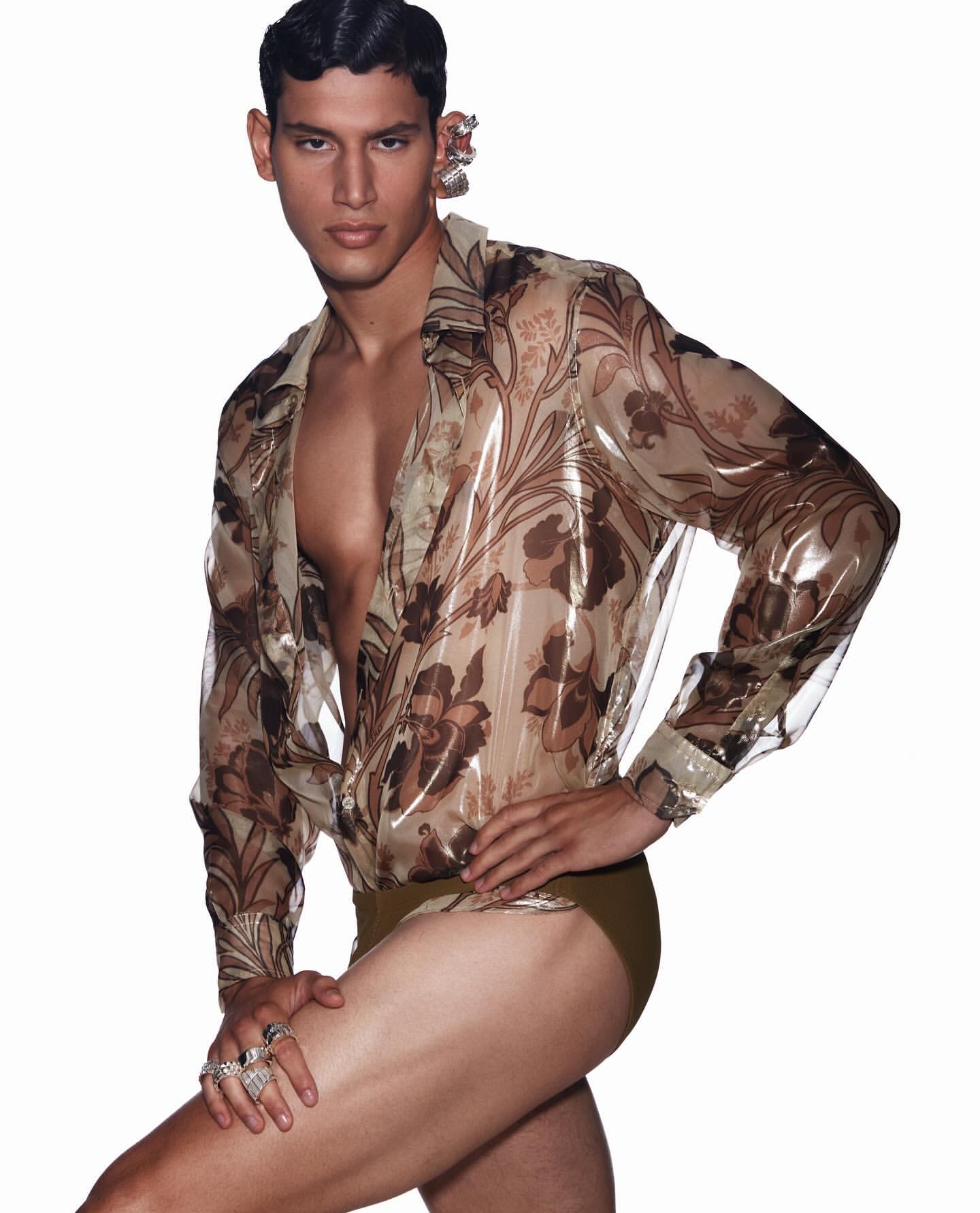 Male model poses in a semi-transparent floral shirt and briefs against a white backdrop, showcasing a fashion-forward and bold style.
