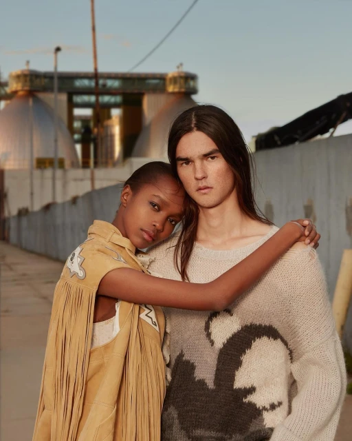 A tan-skinned woman and a light-skinned man with long dark hair pose in an outdoor setting with an industrial background.