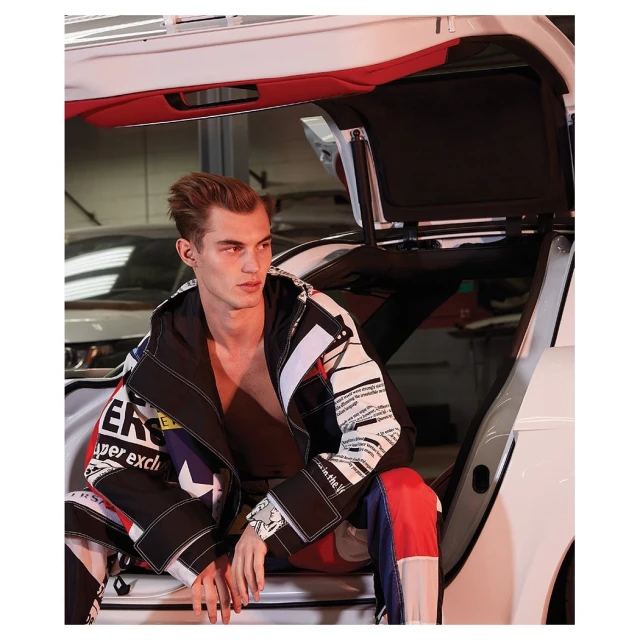 Male model wearing designer jacket sitting in a white car with open doors, photographed indoors with studio lighting and high fashion style.