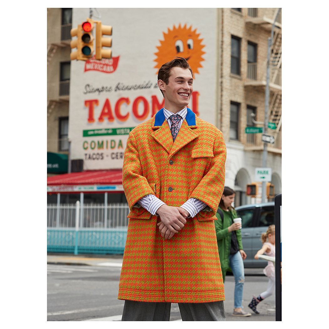 A smiling man in an orange coat stands on a city street with a taco restaurant mural in the background on a bright day.