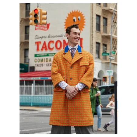 A smiling man in an orange coat stands on a city street with a taco restaurant mural in the background on a bright day.