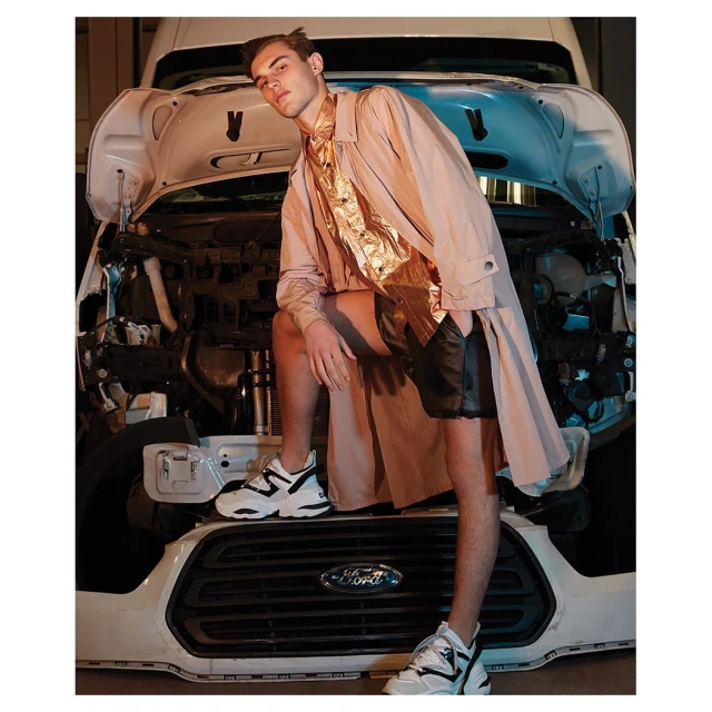 Male model in a beige coat and black shorts poses in front of a white Ford van with the hood open in an indoor setting.