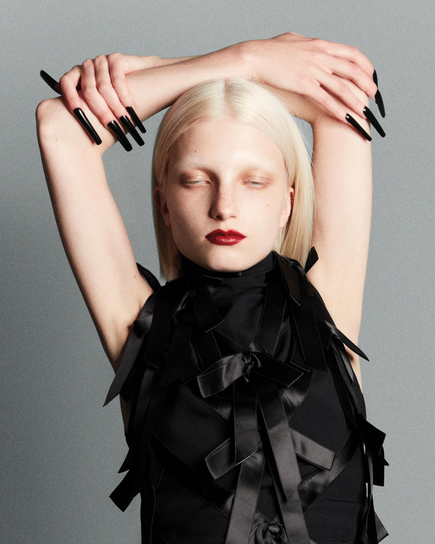 Woman with platinum blonde hair and extra long black nails poses in black dress with bow details in a studio environment.