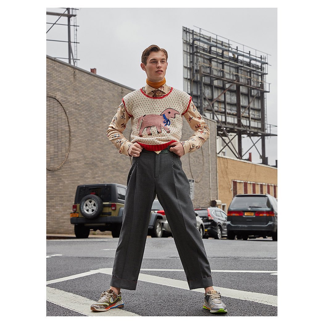 A young adult male poses on a city street wearing an animal print sweater vest, collared shirt, grey slacks, and sneakers with buildings in the background.